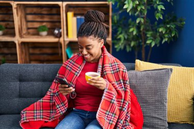 Afro-Amerikalı kadın kahve içiyor ve evde akıllı telefon kullanıyor.