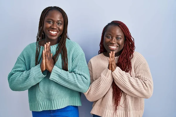 二人のアフリカ人女性が青い背景の上に立ち手を携えて祈りながら自信に満ちた笑顔を許しを求めて — ストック写真