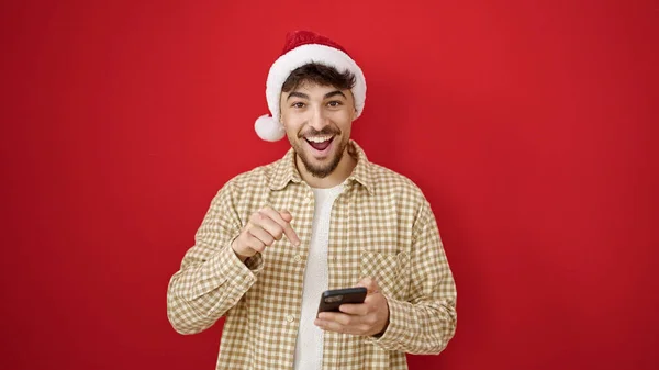 Młody Arabski Człowiek Świątecznym Kapeluszu Wskazując Palcem Smartfona Odizolowanym Czerwonym — Zdjęcie stockowe