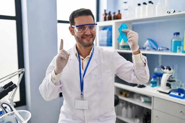 幸福そうな顔をした指を指差すアイデアや質問に驚いた青いリボンを持っている科学研究所で働くひげを持つ若いヒスパニック系の男 ナンバーワン — ストック写真