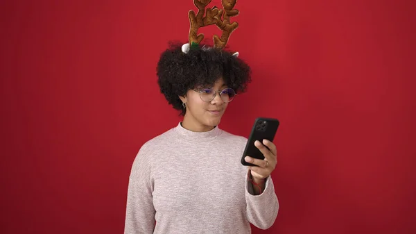 若いですアフリカ系アメリカ人女性身に着けていますクリスマストナカイ耳使用スマートフォン上の隔離された赤の背景 — ストック写真