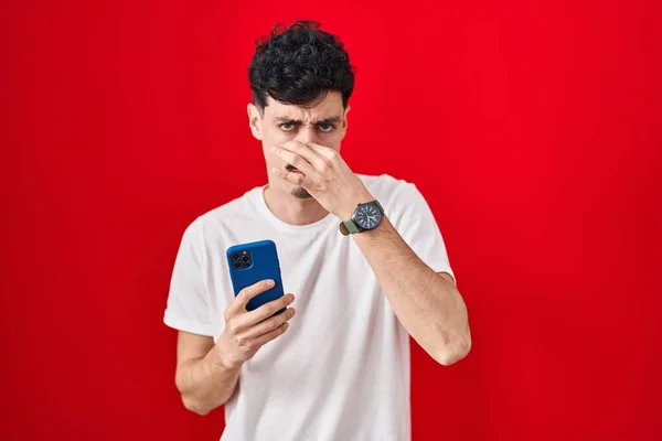 Hispanic Man Using Smartphone Red Background Smelling Something Stinky Disgusting — Stock Photo, Image