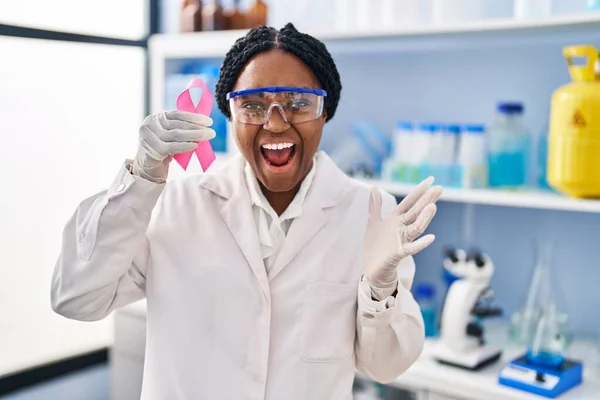 ピンクリボンを手に科学研究所で働くアフリカ系アメリカ人女性が笑顔で勝利を祝い 手を上げて受賞者の表情 — ストック写真