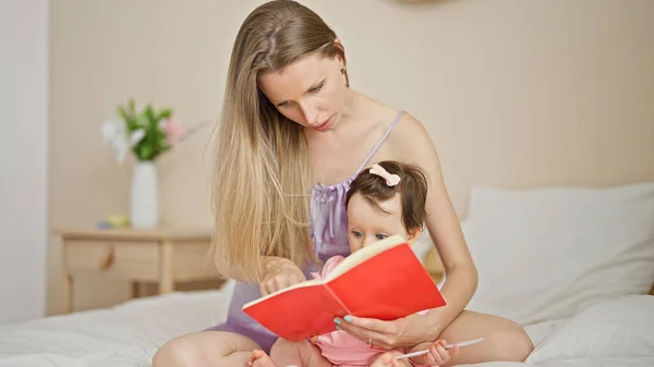 Mother and daughter sitting on bed reading book at bedroom