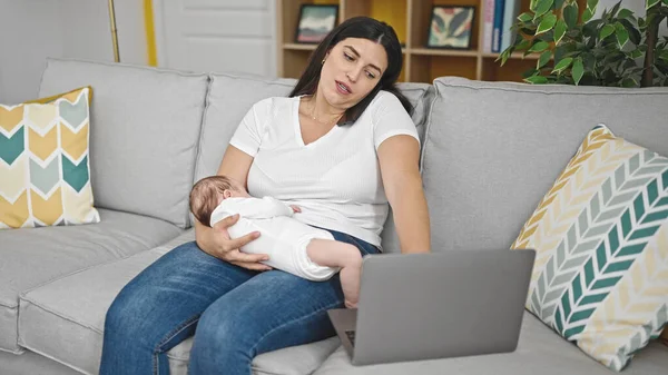 自宅でスマートフォンやラップトップを使用して赤ちゃんを抱えているソファーに座っている母と赤ちゃんの娘 — ストック写真