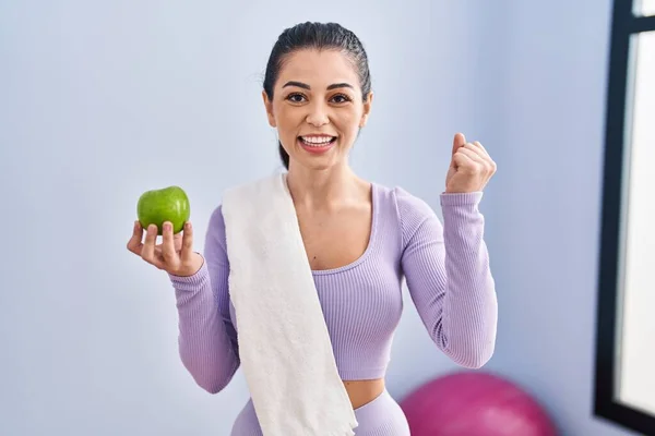 スポーツウェアとタオルを身に着けている若い女性は 誇りに思って叫んで緑のリンゴを食べます 勝利と成功を祝う非常に腕を上げ興奮して — ストック写真