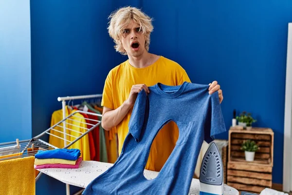 Mladý Blonďatý Muž Drží Tričko Spálenou Dírou Překvapivým Výrazem Prádelně — Stock fotografie