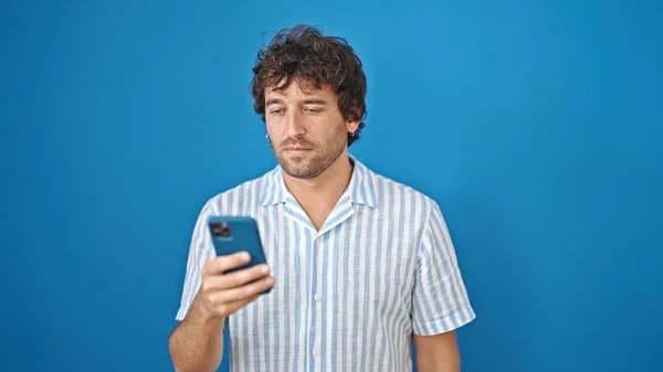 Молодой Латиноамериканец Использует Смартфон Серьезным Выражением Изолированном Синем Фоне — стоковое фото