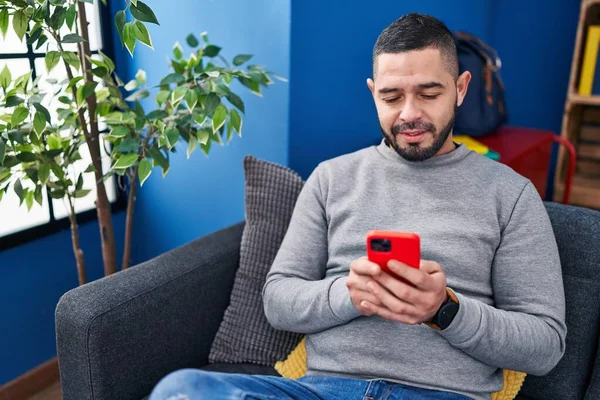 年轻的拉丁男人坐在家里的沙发上用智能手机 — 图库照片