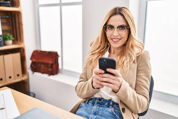 若いブロンド女性ビジネス労働者笑顔自信を持って使用してスマートフォンでオフィス — ストック写真