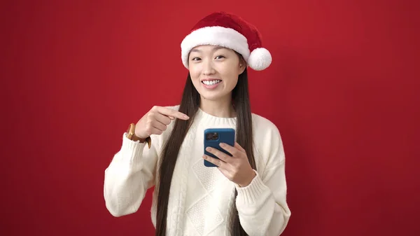 若いです中国の女性身に着けていますクリスマス帽子ポインティングに指でスマートフォン上の隔離された赤の背景 — ストック写真