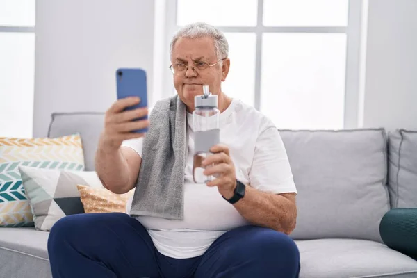 中年白发男人在家里拿着一瓶水 用智能手机制作自拍 — 图库照片