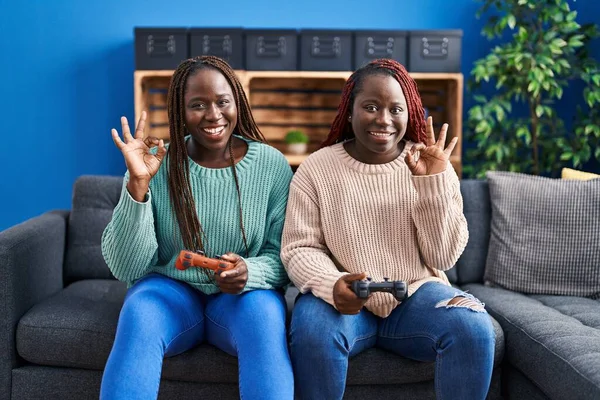 两名玩电子游戏的非洲女人拿着控制器 用手指做着确定的手势 微笑着表示友善的手势 展示着极好的象征 — 图库照片