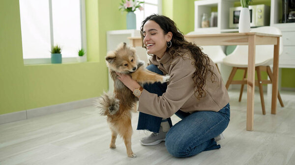 Young hispanic woman with dog sitting on floor hugging at dinning room