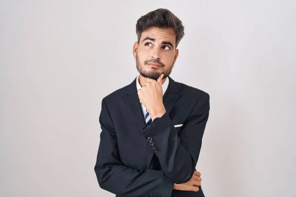 ビジネススーツを着た入れ墨を持つ若いヒスパニック系の男と質問について考える顎に手でネクタイ 集中的な表現 思慮深い顔で笑顔 疑わしい概念 — ストック写真