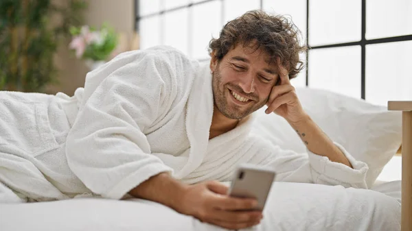 Joven Hombre Hispano Usando Albornoz Usando Smartphone Sonriendo Dormitorio —  Fotos de Stock