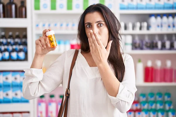 薬局の薬店で若いブルネットの女性が手で口をカバーする丸薬を持って買い物をし ショックを受け 間違いを恐れている 驚きの表情 — ストック写真