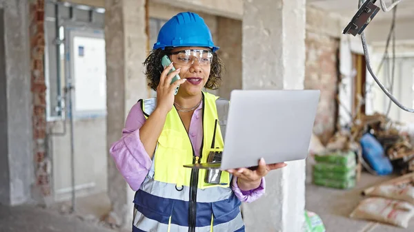 若い美しいラテン語の女性ビルダーは 建設現場でラップトップを使用してスマートフォンで話しています — ストック写真