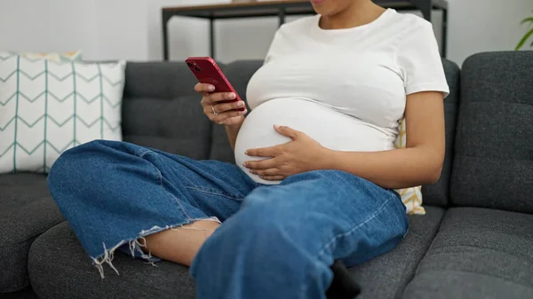 年轻的孕妇坐在家里的沙发上用智能手机 — 图库照片