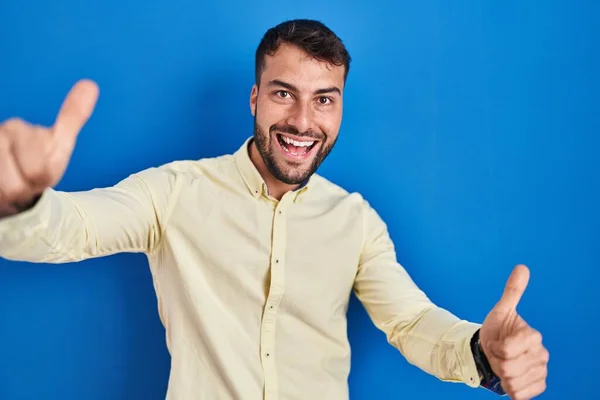 青い背景の上に立つハンサムなヒスパニック系の男性は 手で積極的なジェスチャーを行うことを承認し 親指を上げて笑顔と成功のために幸せ 勝者のジェスチャー — ストック写真