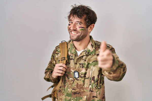 ヒスパニック系の若い男が幸せと面白い顔を持つカメラに指を指してカモフラージュ軍の制服を着て 良いエネルギーと振動は — ストック写真