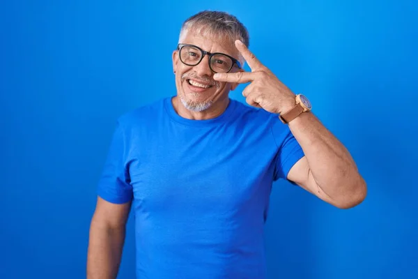 白发苍苍的西班牙裔男子站在蓝色的背景上 用手指捂住脸 愉快地微笑着 表示胜利 — 图库照片