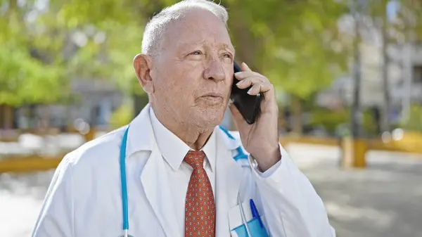 シニア白髪の男医師立っています真剣な表情でスマートフォンで話しています公園で — ストック写真
