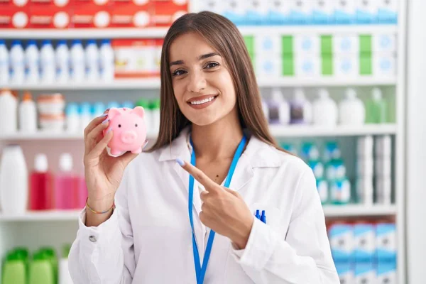 Young Brunette Woman Working Pharmacy Drugstore Holding Piggy Bank Smiling — ストック写真