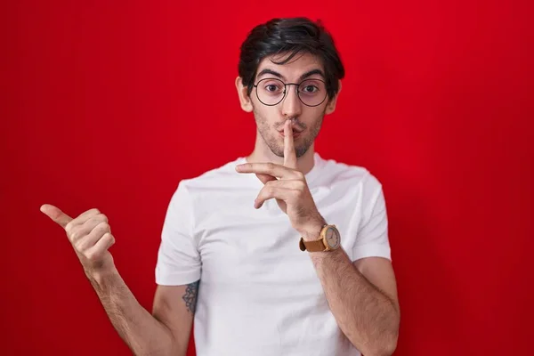 若いヒスパニック系の男性は赤い背景の上に立って唇の側に手を指して上の指で静かにするように求めている 沈黙と秘密の概念 — ストック写真