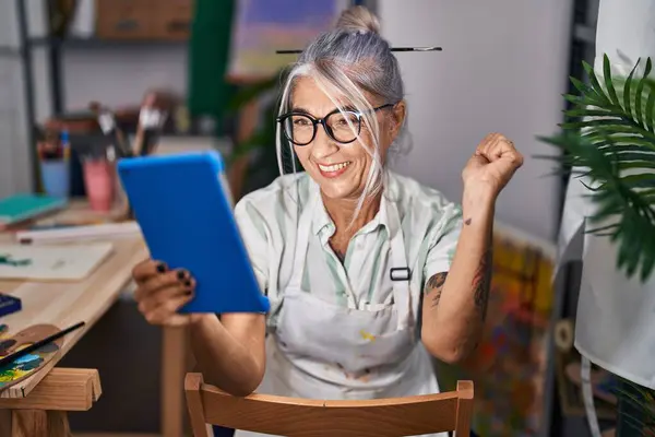 中年妇女灰白的头发坐在艺术工作室里 用平板电脑高喊着自豪 高举双臂庆祝胜利和成功 — 图库照片