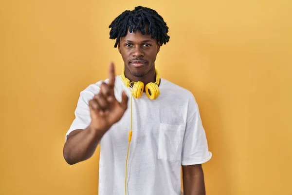指を上げて怒りの表情で指差す黄色い背景の上に立っているドレッドロックを持つ若いアフリカの男は ジェスチャーを示しません — ストック写真