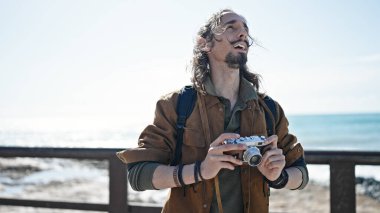 Sırt çantalı genç İspanyol turist deniz kenarında kamera tutuyor.