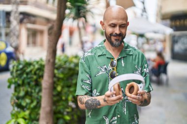 Genç kel adam gülümsüyor. Kulaklığı sokaktan tutuyor.