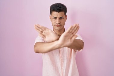 Young hispanic man standing over pink background rejection expression crossing arms and palms doing negative sign, angry face 