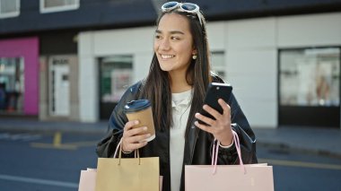 Genç ve güzel İspanyol kadın akıllı telefon kullanıyor. Elinde alışveriş poşetleri ve sokakta kahve tutuyor.