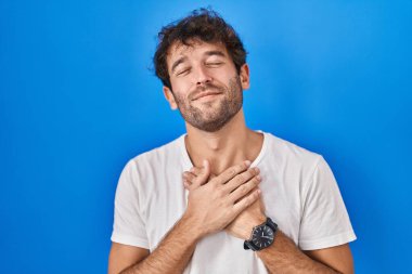 Mavi arka planda duran İspanyol genç adam elleri göğsünde, gözleri kapalı ve yüzünde minnet dolu bir jest ile gülümsüyor. sağlık kavramı. 