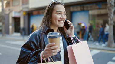 Güzel İspanyol bir kadın akıllı telefondan konuşuyor. Elinde alışveriş poşetleri ve sokakta kahve var.