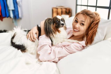 Beyaz bir kadın yatakta uzanıyor. Yatak odasında köpeğe sarılıyor.