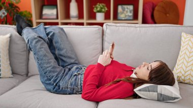 Evdeki kanepede uzanmış akıllı telefon kullanan genç İspanyol kadın.