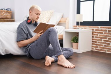 Genç beyaz adam kitap okuyor. Yatak odasında yerde oturuyor.