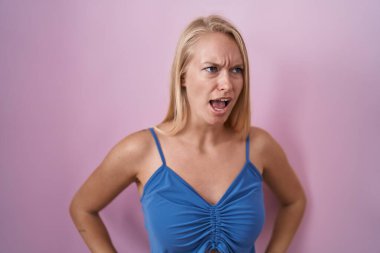 Young caucasian woman standing over pink background angry and mad screaming frustrated and furious, shouting with anger. rage and aggressive concept. 