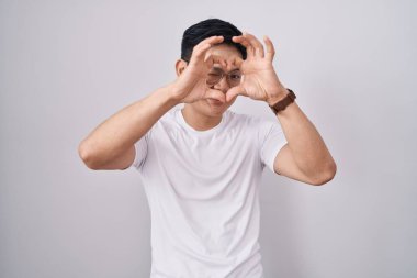 Beyaz arka planda duran genç Asyalı adam kalp şeklinde elleriyle ve parmaklarıyla tabelanın içinden bakıp gülümsüyor. 