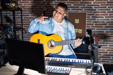 Latin asıllı genç adam müzik stüdyosunda klasik gitar çalıyor neşeli bir şekilde gülümsüyor ve parmaklarını ve ağzını işaret ediyor. diş sağlığı kavramı. 