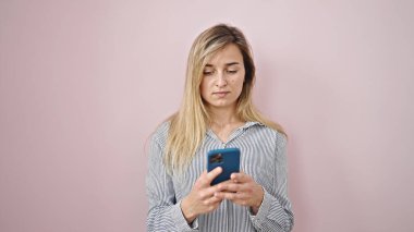 Soyutlanmış pembe arka planda ciddi bir ifadeyle akıllı telefon kullanan genç sarışın kadın.