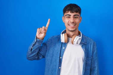 Young hispanic man standing over blue background showing and pointing up with finger number one while smiling confident and happy. 