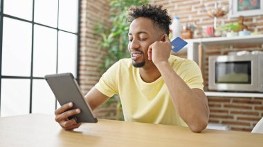 Afro-Amerikalı bir adam dokunmatik ped ve kredi kartıyla alışveriş yapıyor yemek odasındaki kanepede oturuyor.