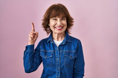 Pembe arka planda duran orta yaşlı bir kadın bir numarayı gösterip işaret ederken kendinden emin ve mutlu bir şekilde gülümsüyor.. 