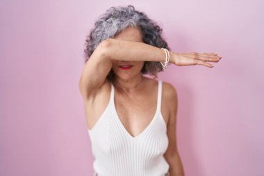 Pembe arka planda gri saçlı orta yaşlı bir kadın gözlerini kollarıyla kapatıyor, ciddi ve üzgün görünüyor. Görme, saklama ve reddetme konsepti 