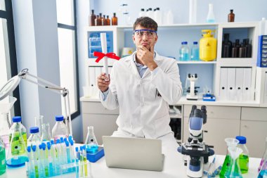 Laboratuvarda çalışan Latin asıllı genç bir adam diplomasını elinde tutarak ciddi bir yüz ifadesiyle eli çenesinde soru soran, kafa karıştırıcı bir fikir hakkında düşünceli biri. 