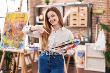 Sanat stüdyosundaki genç beyaz kadın palet tutarak mutlu ve gururlu bir kendini işaret ediyor. 